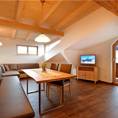 Ein modernes Wohnzimmer mit einem Esstisch und einer gemütlichen Sitzecke. Große Fenster sorgen für viel Tageslicht und eine einladende Atmosphäre.