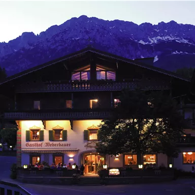 A traditional inn in the mountains, surrounded by majestic peaks. The lighting of the house radiates an inviting atmosphere.