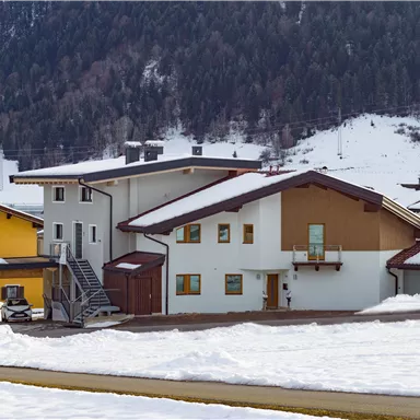Ein Winterdorf mit mehreren Häusern, umgeben von Schnee und Wald. Die Gebäude haben verschiedene Farben und architektonische Stile.