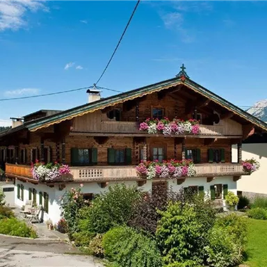 A traditional wooden house in the Alps with blooming plants on the balcony. The surroundings are green and the sky is blue.