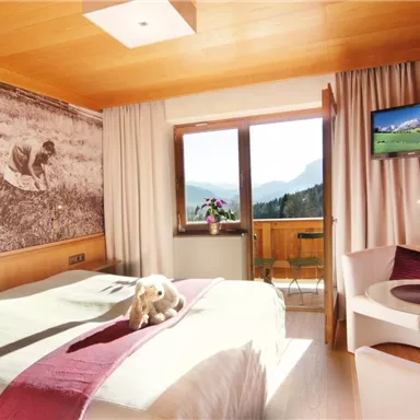 A bright hotel room with a double bed and wooden walls. A mountain landscape can be seen through the window.
