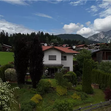 Eine ruhige Wohngegend mit mehreren Häusern und üppiger Vegetation. Im Hintergrund sind Berge und ein bewölkter Himmel zu sehen.
