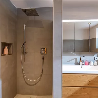 A modern bathroom with a shower and a large wooden bathtub. The walls are designed in neutral gray tones and there is a large window front for natural light.