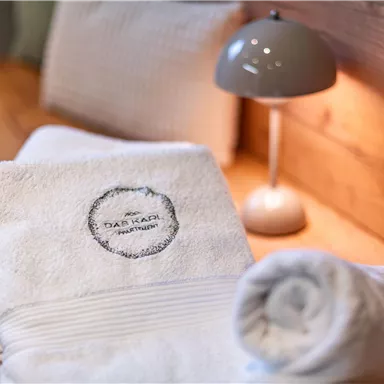 A soft, white stack of towels with a logo. In the background, a table lamp is on a wooden shelf.