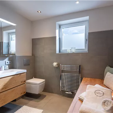 A modern bathroom with wooden cabinets and a window light source. The walls are in soft shades of gray.