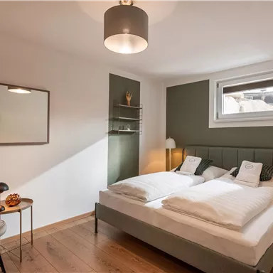 A modern bedroom with a double bed and minimalist decor. The light is bright and friendly, with a cozy side table and a lamp.