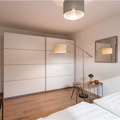 A modern bedroom with a large white wardrobe and a wooden floor. There is a floor lamp and a mirror on the wall.