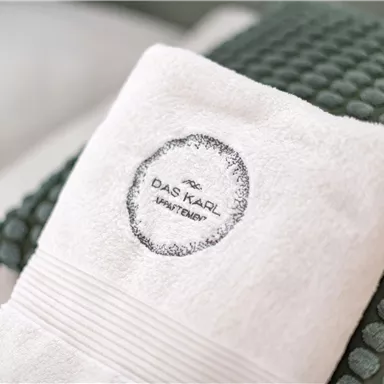 A white towel with the logo of "Das Kaiser Wasser" lies on a green pillow. The background is softly and brightly colored.