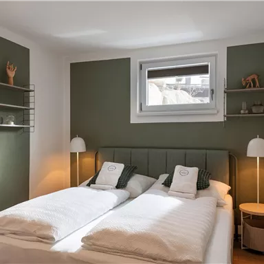 A modern bedroom with two beds and minimalist design. The walls are painted in soft shades of green and there are bedside tables with lamps.