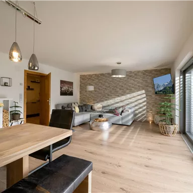 A modern living room with a large window front and a dining area. The stylish furnishings include a comfortable couch and decorative plants.