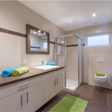 Ein modernes Badezimmer mit Dusche, einem großen Spiegel und einem Waschbecken. Frische Handtücher in leuchtenden Farben sind auf der Ablage und dem Boden platziert.