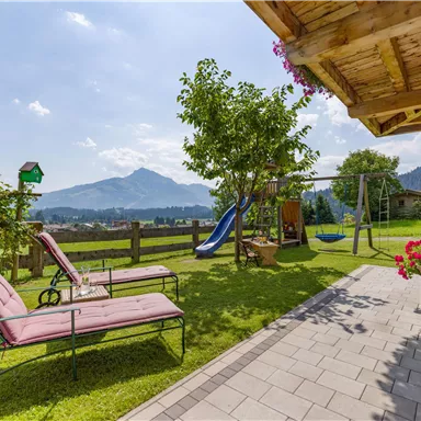 Ein einladender Garten mit Liegen und einem Spielplatz. Im Hintergrund erheben sich Berge unter einem klaren Himmel.