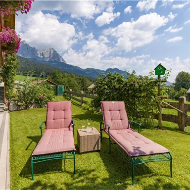 Eine entspannte Gartenlandschaft mit zwei Liegen und einem kleinen Tisch. Im Hintergrund sind grüne Wiesen und Berge unter einem blauen Himmel zu sehen.