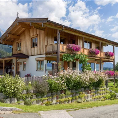 Ein gemütliches Holzhaus mit Balkonen, umgeben von bunten Blumen. Im Hintergrund sind grüne Wiesen und Berge sichtbar.