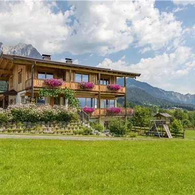 Ein schönes,holzhaus mit Blumenbalkonen in einer grünen Wiesenlandschaft. Im Hintergrund sind Berge und ein blauer Himmel zu sehen.