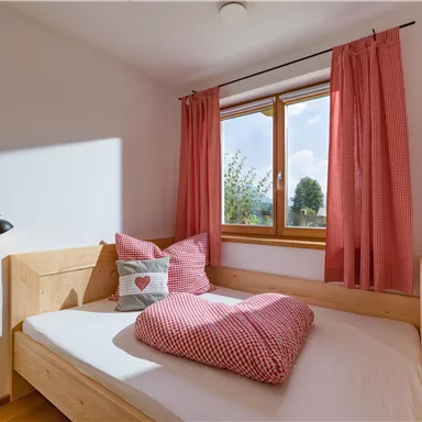 Ein gemütliches Schlafzimmer mit einem Holzbett und roten Vorhängen. Auf dem Bett liegen Kissen mit einem Herzmotiv.