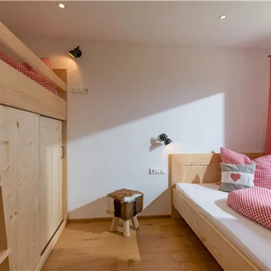 Ein gemütliches Schlafzimmer mit einem Hochbett und einem Einzelbett. Es hat eine warm-holzige Einrichtung und rote Kissen mit einem herzlichen Muster.