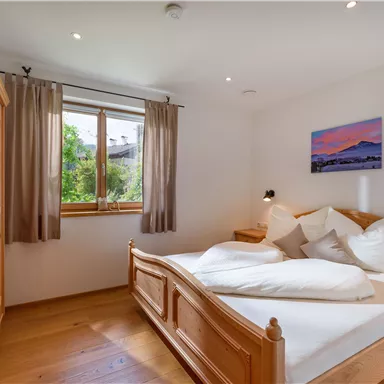 Ein gemütliches Schlafzimmer mit einem großen Bett und Holzmöbeln. Durch das Fenster fällt Tageslicht und blickt auf eine grüne Landschaft.
