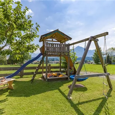 Ein schöner Spielplatz mit einer Rutsche, einer Schaukel und einem Spielhaus. Im Hintergrund sind grüne Wiesen und ein blauer Himmel zu sehen.