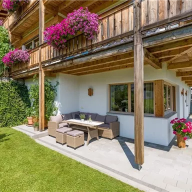 Ein gemütlicher Außenbereich mit einem Tisch und Stühlen unter einem Holzüberhang. Der Bereich ist umgeben von grünem Rasen und blühenden Pflanzen.