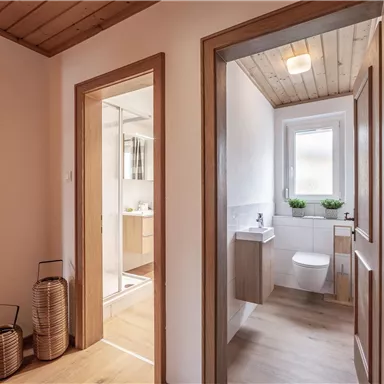 A modern designed corridor with wooden ceilings and bright walls. In the background, a bathroom with a window and modern sanitary facilities is visible.