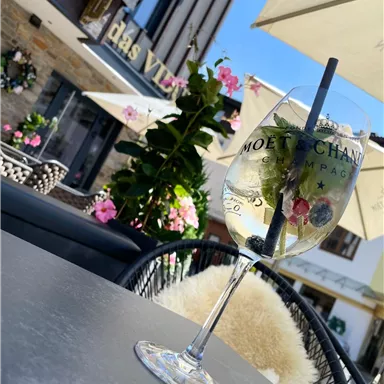 Ein erfrischendes Getränk in einem Glas, dekoriert mit Beeren und einer Limettenscheibe. Im Hintergrund sind blühende Pflanzen und ein Restaurant zu sehen.