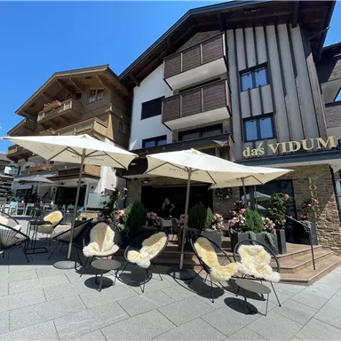 Ein modernes Café mit gemütlichen Sitzplätzen und Sonnenschirmen. Im Hintergrund sieht man ein traditionelles Gebäude mit Holzverkleidung.