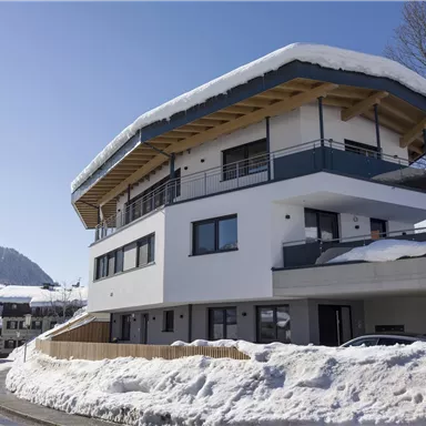Ein modernes Haus mit einem schneebedeckten Dach in einer winterlichen Landschaft. Die Umgebung zeigt eine ruhige Straße und verschneite Berge im Hintergrund.