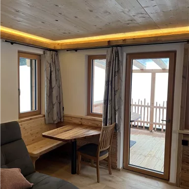 A cozy room with a wooden ceiling and large windows. A table with a chair is next to a comfortable sofa.