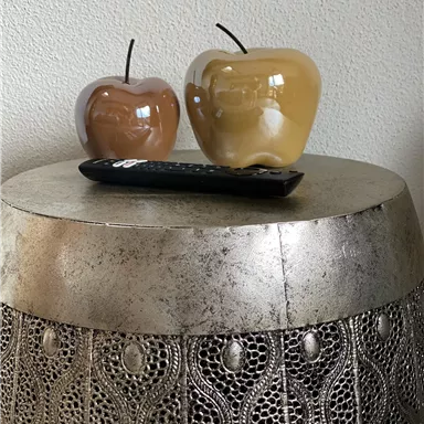 Zwei dekorative Äpfel aus Glas stehen auf einem Tisch. Neben ihnen liegt eine Fernbedienung auf der Tischplatte.