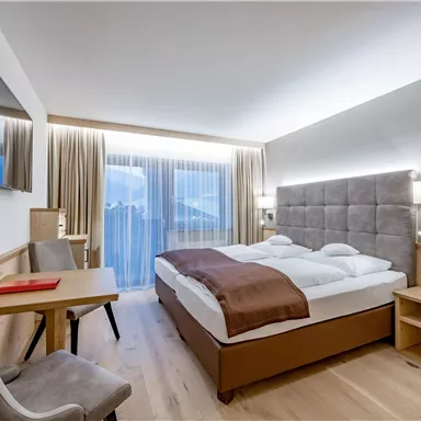 A modern hotel room with a double bed and elegant decor. Large windows provide plenty of daylight and a view outside.