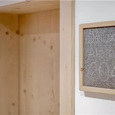 An interior with a wooden frame that displays a sign reading "Deluxe Alpin 214." The wall is light, and the wooden elements give the room a cozy atmosphere.