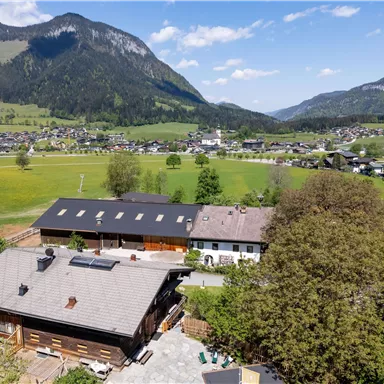 A rural view with traditional houses in the foreground and green fields as well as mountains in the background. The sky is clear and bright blue.