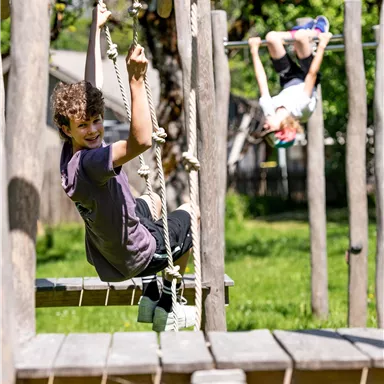 Two children are playing on a playground. One is swinging on a rope, while the other is hanging upside down from the bars.
