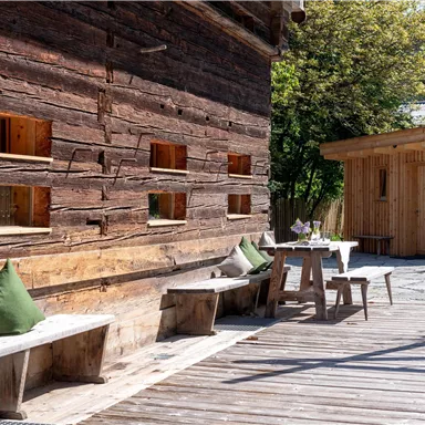 A rustic wooden wall with windows and a walkway. There is a wooden bench and a table with chairs outside.