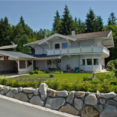 A modern house with a balcony and a surrounding garden. Surrounded by trees and a stone driveway.