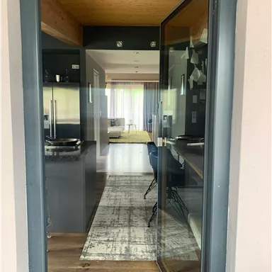 Ein einladender Eingang zu einem modernen Raum mit Holzbalkendecke. Im Hintergrund sieht man einen hellen Wohnbereich mit Fenster und eleganten Möbeln.