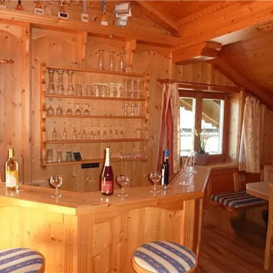Eine gemütliche Bar aus Holz mit eleganten Gläsern und mehreren Flaschen Wein. Im Hintergrund befinden sich ein Fenster und eine Sitzecke mit bequemen Stühlen.