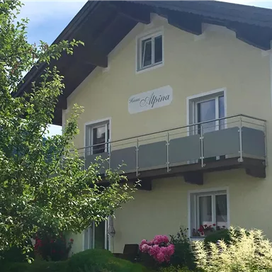 A charming house with a balcony and a clear sky. Surrounded by green trees and colorful flowers.