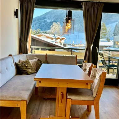 A cozy dining area with a wooden infotable and comfortable benches. Through large windows, one looks out at snow-covered mountains and a tranquil landscape.