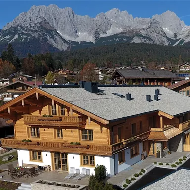 A cozy wooden house with a large balcony, surrounded by breathtaking mountain landscapes. The sun shines over the picturesque surroundings.