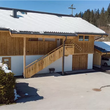 A modern building with wooden cladding and a snow-covered roof. On the right side, there are parking spaces and a path with a staircase.