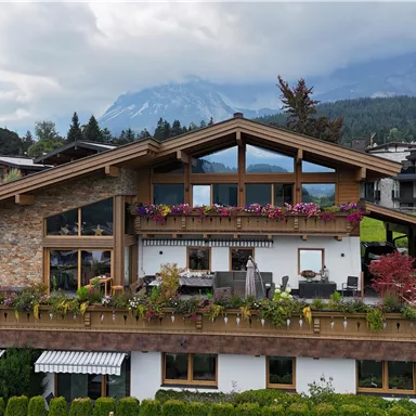 A modern house in the mountains with wooden cladding and colorful flowers. In the background are majestic mountains and a cloudy sky.