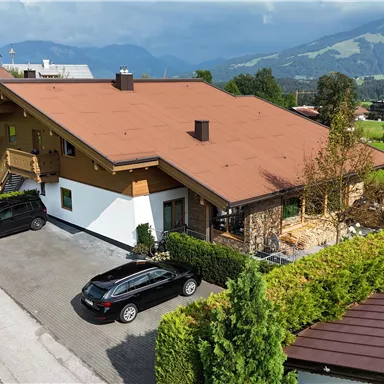 A modern house with a red roof in a quiet residential area.  
Surrounded by green scenery and trees, several cars are parked in the driveway.