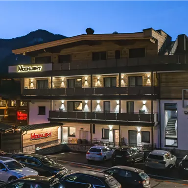 Ein modernes Hotelgebäude bei Nacht, umgeben von Bergen. Parkplätze sind direkt davor sichtbar.