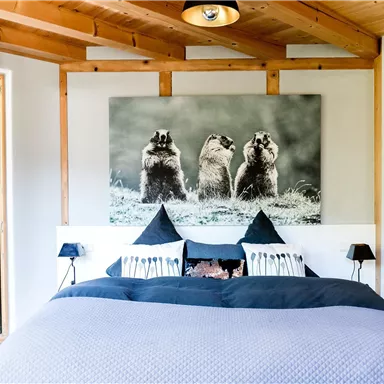 Ein helles Schlafzimmer mit einem großen Bett und Holzdecke. An der Wand hängt ein Bild von drei Tieren, und es gibt viel natürliches Licht.
