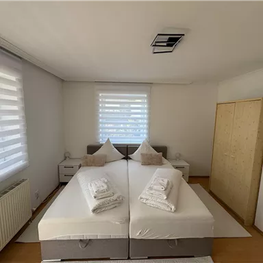 A bright bedroom with two single beds and fresh towels. Large windows let in plenty of daylight.