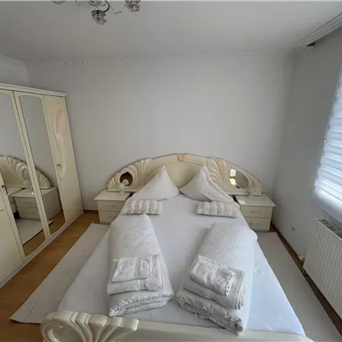 A bright bedroom with a large bed and beige walls. There is a mirror and a large wardrobe in the room.