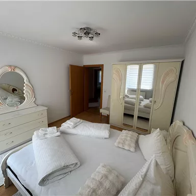 A bright bedroom with a large bed and an elegant dresser. The room is simply and modernly furnished, with a large mirror and a spacious wardrobe.