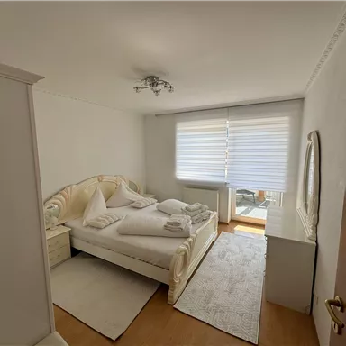 A bright bedroom with a large bed and white bedding. There is a large window with blinds and a mirror on the wall.